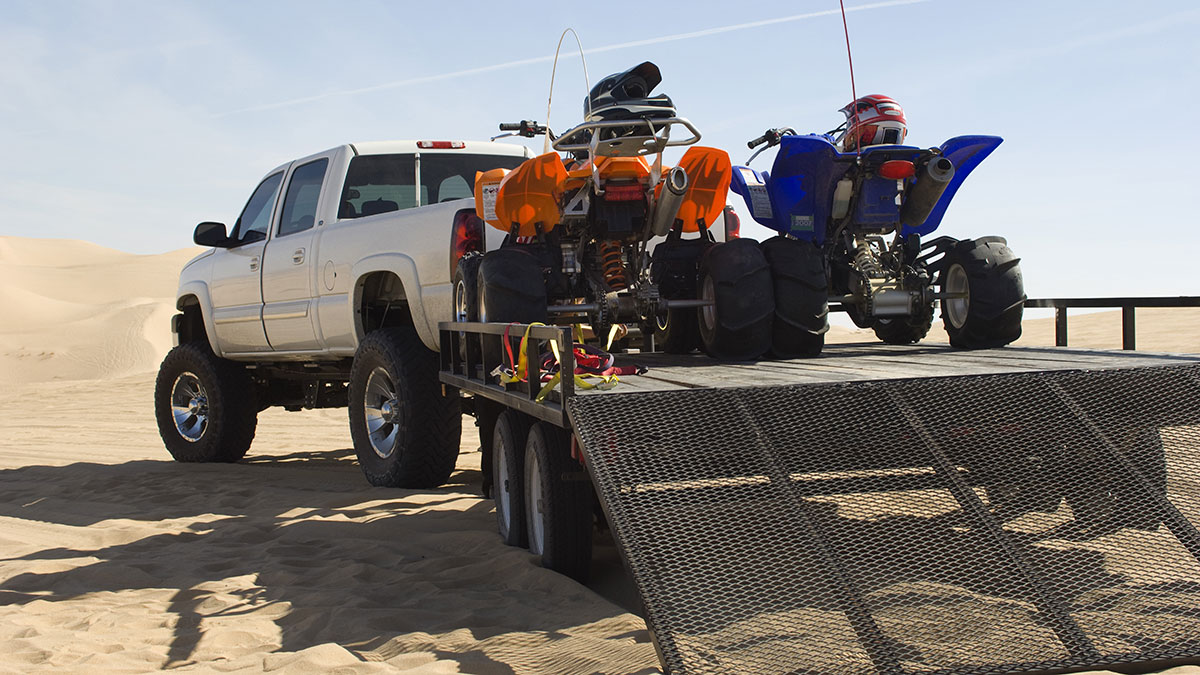 Two ATVs loaded on a trailer attached to a large white pickup truck parked amoung sand dunes.